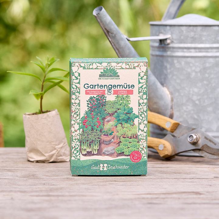 Paquete de semillas de hortalizas de jardín sobre una mesa de madera, junto a una pequeña planta, una regadera y tijeras de podar en el fondo.