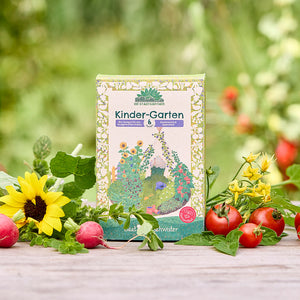 Un paquete de semillas orgánicas “Kinder-Garten” se encuentra sobre una mesa de madera, rodeado de girasoles, rábanos, tomates y vegetación fresca del jardín.