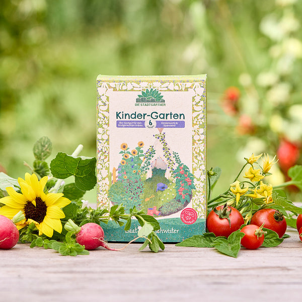 Un paquete de semillas orgánicas “Kinder-Garten” se encuentra sobre una mesa de madera, rodeado de girasoles, rábanos, tomates y vegetación fresca del jardín.
