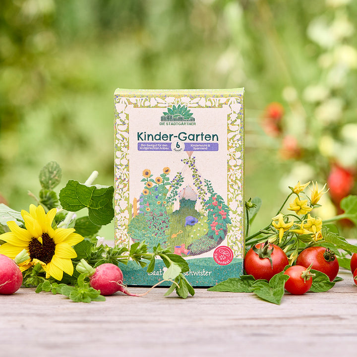 Un paquete de semillas orgánicas “Kinder-Garten” se encuentra sobre una mesa de madera, rodeado de girasoles, rábanos, tomates y vegetación fresca del jardín.