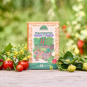 Un paquete de semillas de "Naschgarten" se encuentra sobre una mesa de madera en el jardín, rodeado de tomates frescos, fresas y follaje verde.