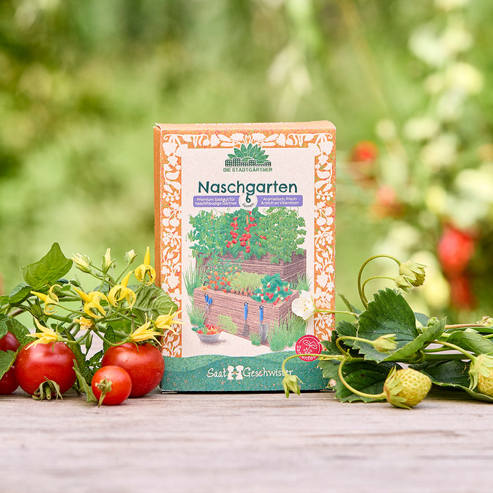 Un paquete de semillas de "Naschgarten" se encuentra sobre una mesa de madera en el jardín, rodeado de tomates frescos, fresas y follaje verde.