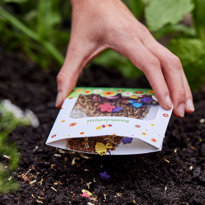 Una mano esparce semillas de flores desde una bolsa decorada de colores sobre la tierra del jardín.