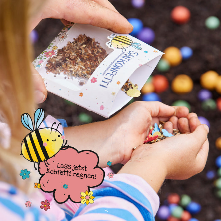 Un niño sostiene confeti de semillas en las manos mientras le vierten encima un paquete de semillas con motivos florales. En primer plano, se muestra el dibujo de una abeja con el bocadillo "¡Que llueva confeti!". Al fondo, bolas de colores sobre tierra.