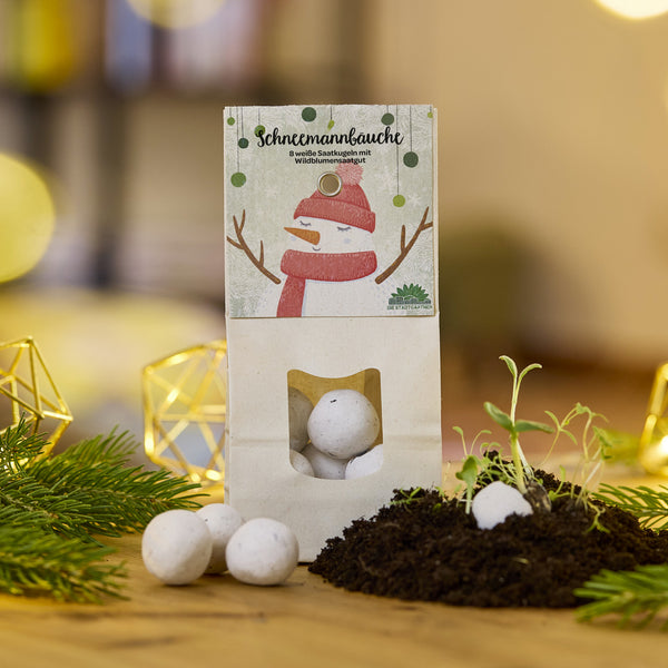 Un paquete de bolas de semillas blancas llamadas "Snowman Bellies" se encuentra sobre una mesa, al lado de tierra, plántulas y ramas de abeto.