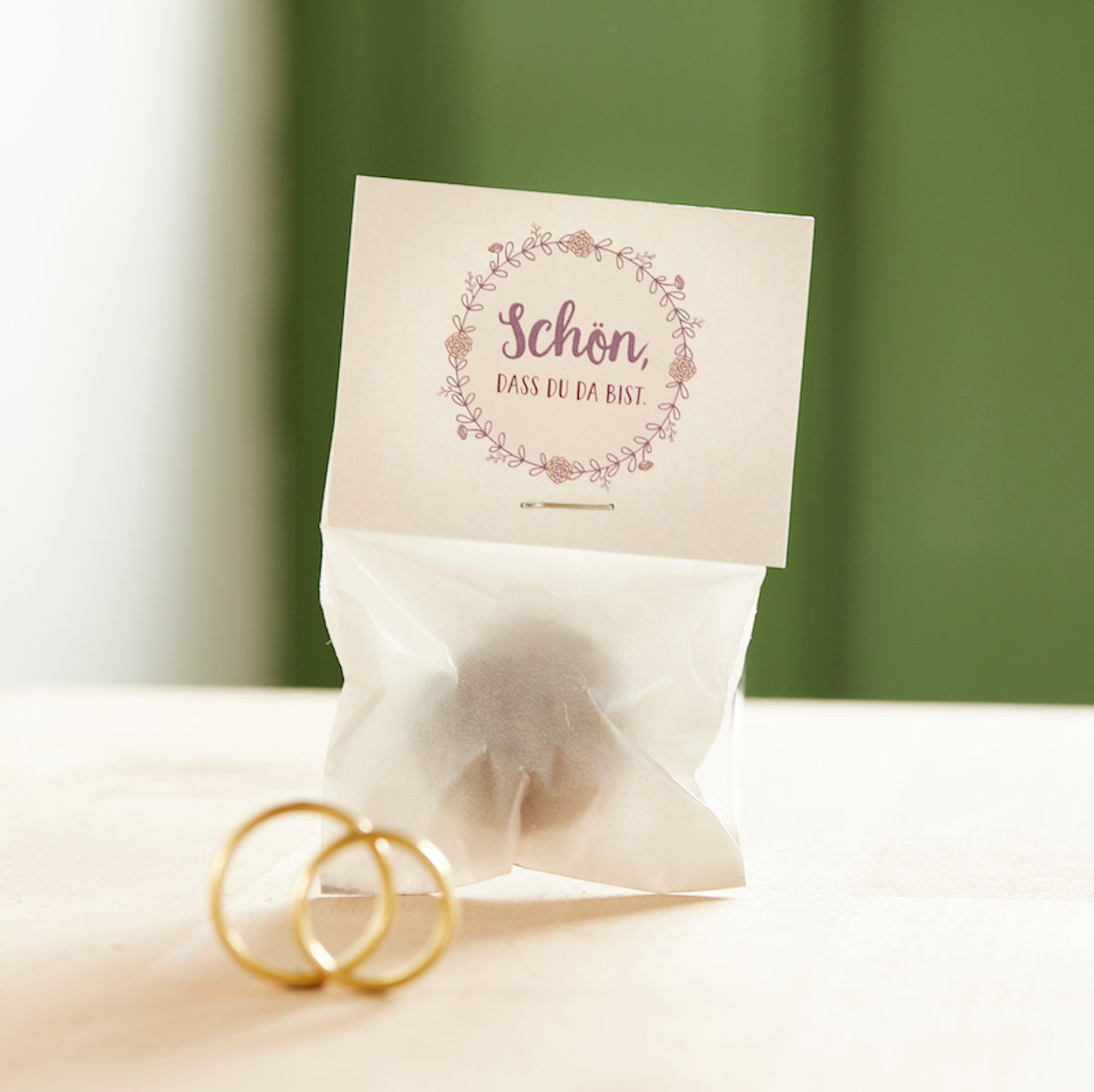 Pequeña bolsa con la inscripción "Es genial que estés aquí" y dos anillos de boda de oro delante.