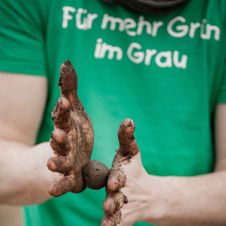 Una persona con una camiseta verde con la inscripción "Por más verde en el gris" muestra las manos sucias con tierra.