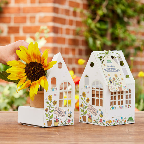 Mano con girasol y dos paquetes decorativos en forma de casa con la inscripción "Jardín de flores" sobre una mesa de madera al aire libre.