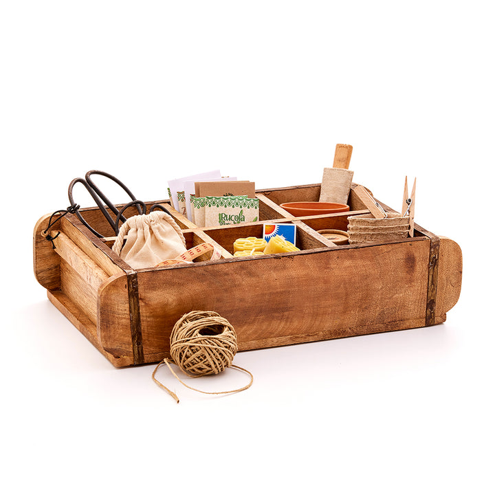 Caja de madera con diversos utensilios de jardinería como tijeras, bolsas de semillas, macetas, cordel y otros accesorios.