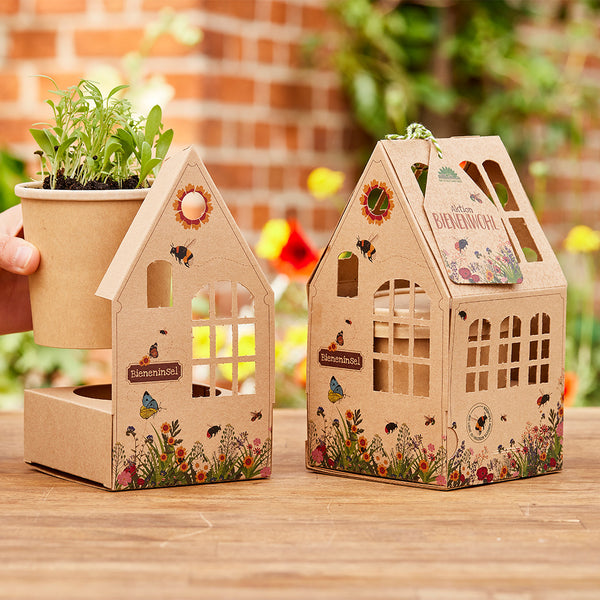 Dos casas decorativas de cartón con motivos de flores e insectos, una de ellas con una maceta que contiene plántulas verdes, ambas colocadas sobre una mesa de madera en el jardín.