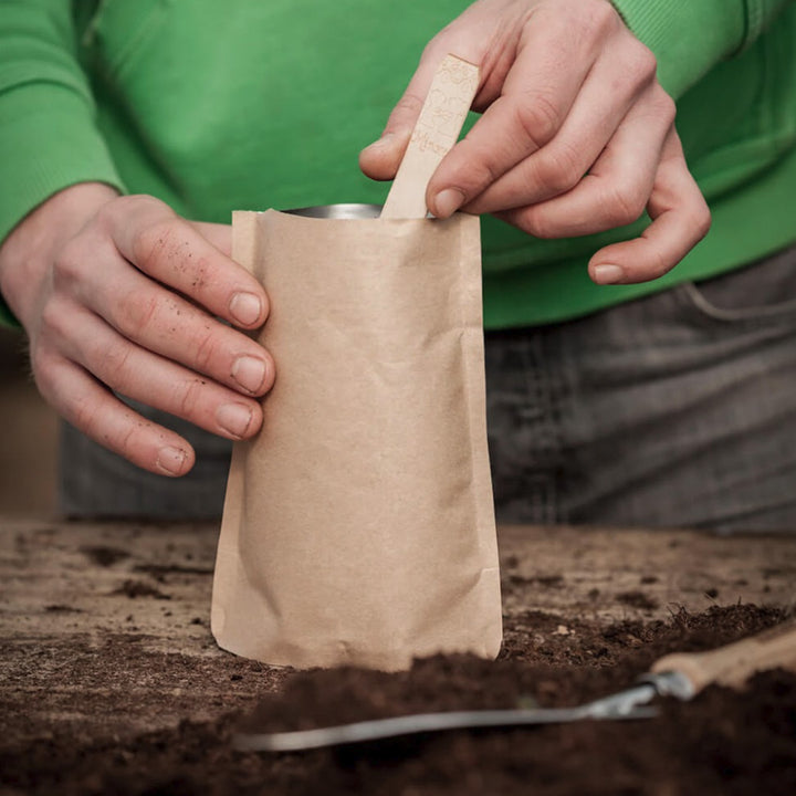 Dos manos sostienen una bolsa de papel marrón, mientras una mano inserta un palo de madera en ella; en primer plano, sobre una mesa de madera hay tierra y una herramienta de jardinería.