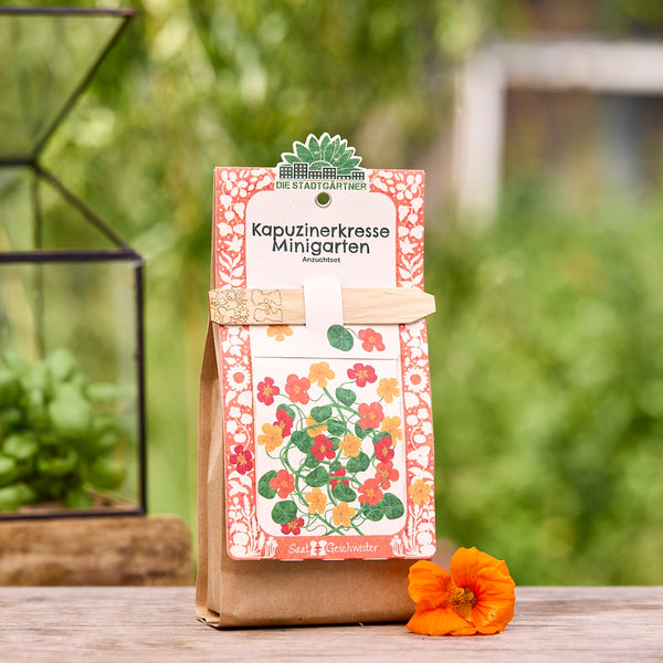 Bolsa marrón que contiene un kit de cultivo de capuchina, decorado con una colorida ilustración de las flores y un tutor de madera, en el jardín sobre una mesa de madera, junto a una flor de capuchina naranja.