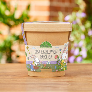 Taza marrón con una banderilla colorida que representa “Flores de Pascua en una taza” y dos conejitos de Pascua dibujados con flores, de pie sobre una superficie de madera en el jardín.