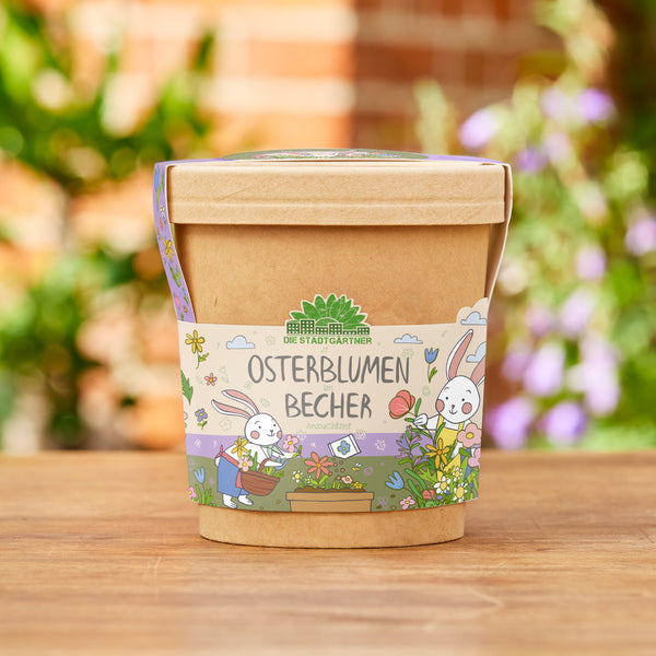 Taza marrón con una banderilla colorida que representa “Flores de Pascua en una taza” y dos conejitos de Pascua dibujados con flores, de pie sobre una superficie de madera en el jardín.