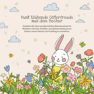 Ilustración de un conejo blanco en un prado colorido con flores primaverales, mariposas y nubes; en la parte superior hay un texto en alemán sobre la alegría de Pascua y la decoración floral.
