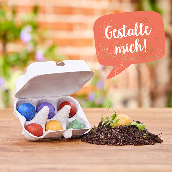 Un cartón de huevos abierto con bombas de semillas de colores y un montículo de tierra con plántulas sobre una mesa de madera; un bocadillo de diálogo con el texto "¡Diséñame!"
