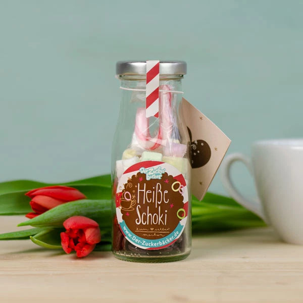 Botella con etiqueta “Chocolate Caliente” y pajita, junto a un tulipán rojo y una taza al fondo.