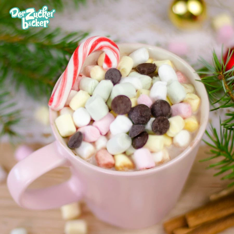 Una taza de chocolate caliente con mini malvaviscos y chispas de chocolate, junto a un bastón de caramelo, rodeado de ramas de abeto y adornos navideños.
