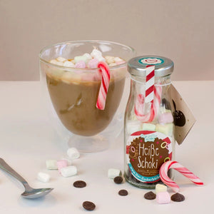 Un vaso de chocolate caliente con malvaviscos, al lado una botella con la etiqueta "Chocolate caliente" y bastones de caramelo, con malvaviscos y chispas de chocolate esparcidas en primer plano.