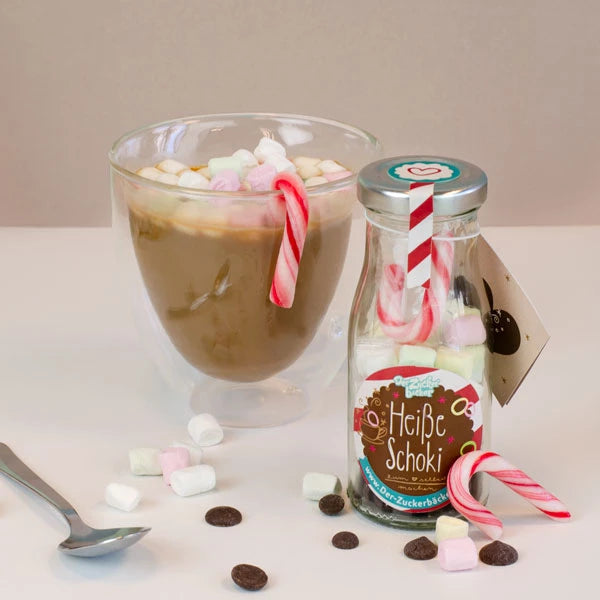 Un vaso de chocolate caliente con malvaviscos, al lado una botella con la etiqueta "Chocolate caliente" y bastones de caramelo, con malvaviscos y chispas de chocolate esparcidas en primer plano.