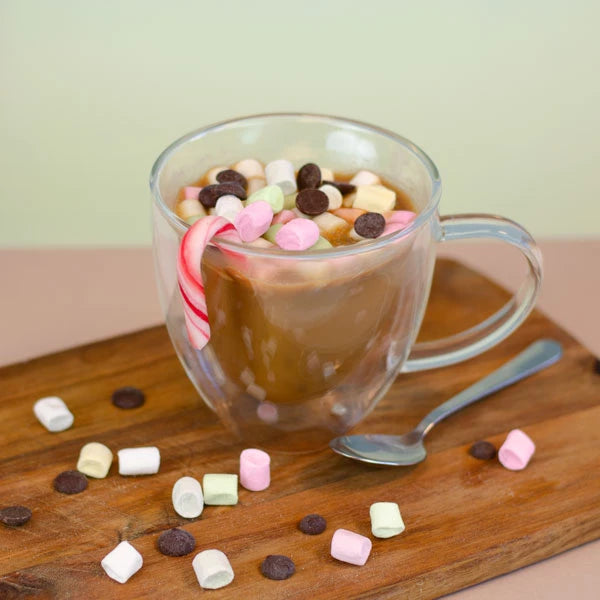 Chocolate caliente en taza transparente con malvaviscos, chispas de chocolate y bastones de caramelo, servido en una tabla de madera.