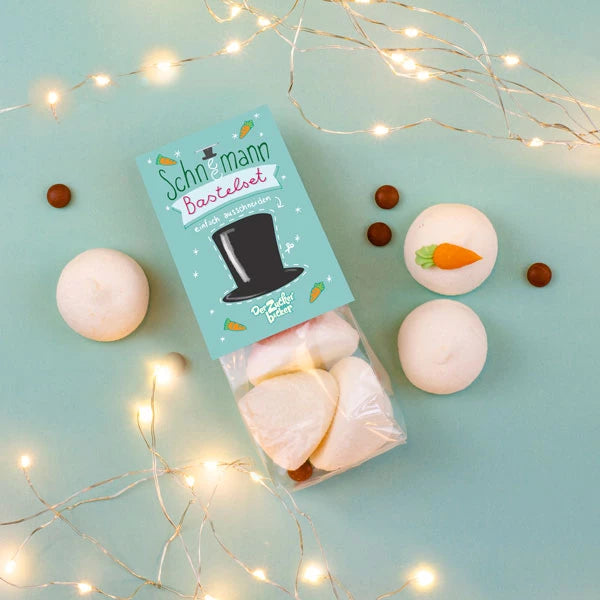 Embalaje con nubes "kit muñeco de nieve", rodeadas de pequeñas bolas de chocolate y luces de colores sobre un fondo verde menta.
