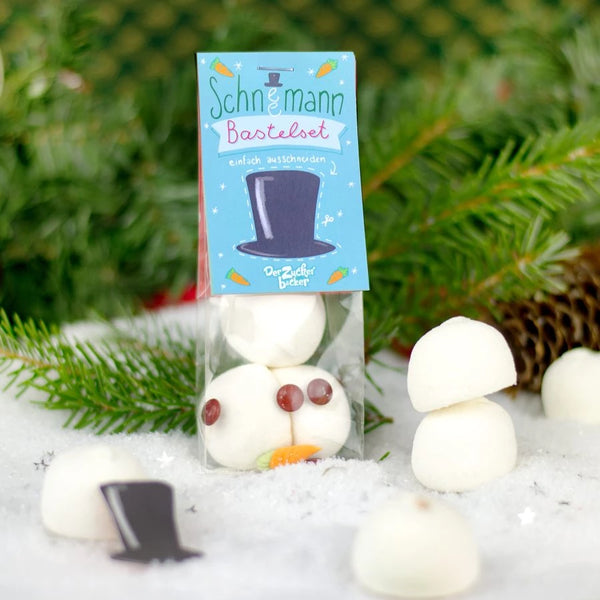 Kit de manualidades de muñeco de nieve empaquetado con malvaviscos y elementos decorativos sobre un lecho de nieve con verde abeto de fondo.