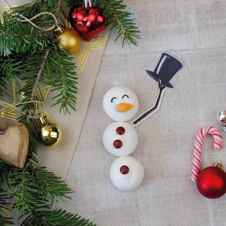 Un muñeco de nieve hecho de malvaviscos con un pan de azúcar, gotas de chocolate y una nariz de zanahoria yace sobre una tela junto a adornos navideños y ramas de abeto.