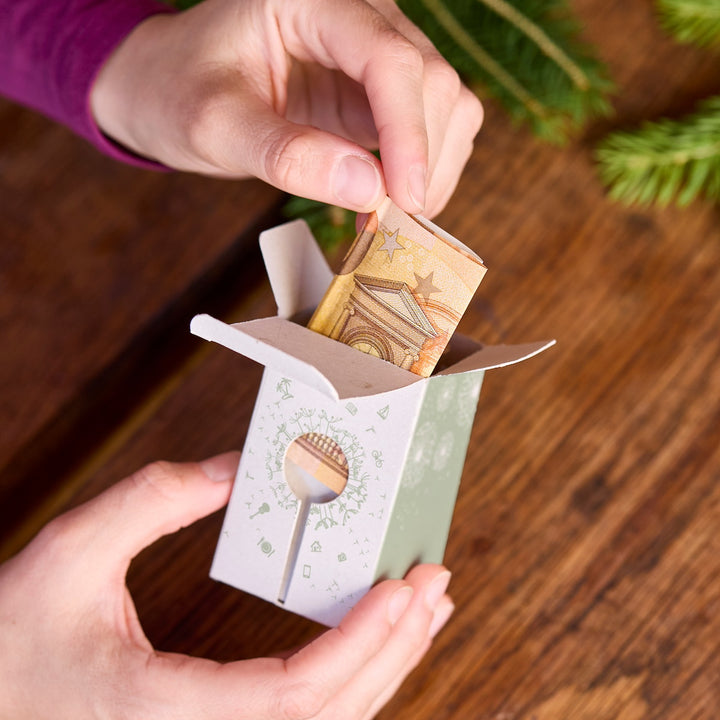 Unas manos colocan un billete de euro doblado dentro de una pequeña caja de regalo de papel sobre una mesa de madera.