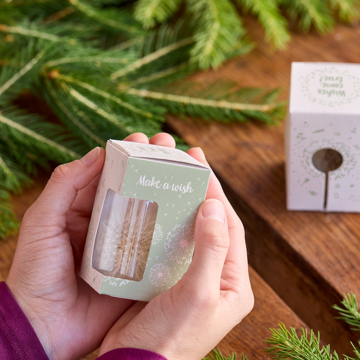 Manos sosteniendo una pequeña caja de regalo con la inscripción "Pide un deseo" junto a ramas de abeto en una mesa de madera.