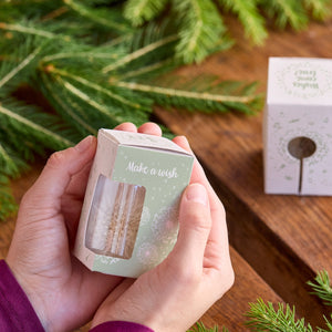 Manos sosteniendo una pequeña caja de regalo con la inscripción "Pide un deseo" junto a ramas de abeto en una mesa de madera.