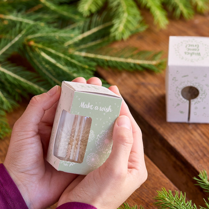 Manos sosteniendo una pequeña caja de regalo con la inscripción "Pide un deseo" junto a ramas de abeto en una mesa de madera.