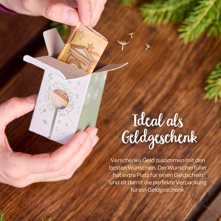 Unas manos colocan un billete doblado en una pequeña caja de regalo de papel, con el texto "Ideal como regalo en efectivo" escrito al lado sobre una mesa de madera con ramas de abeto.