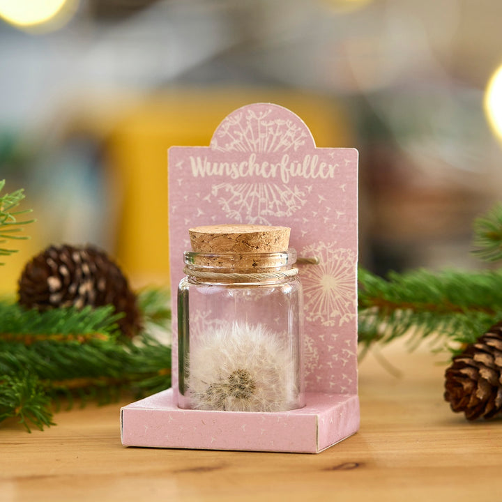 Un pequeño frasco de vidrio con corcho, que contiene un diente de león, se encuentra sobre un paquete rosa con la inscripción "Cumplidor de deseos", decorado con ramas de abeto y piñas.