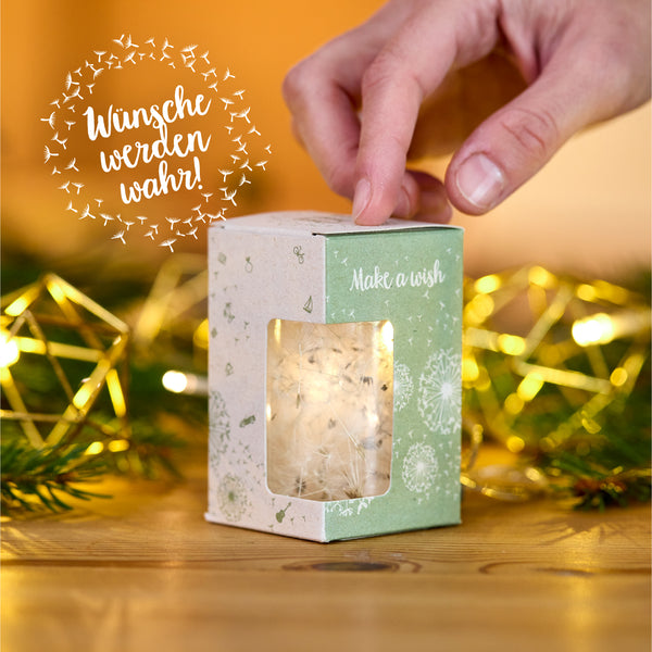 Mano sosteniendo una caja de regalo con sombrillas de diente de león y la inscripción "Pide un deseo", en el fondo luces y ramas de abeto, junto a la inscripción "¡Los deseos se hacen realidad!".