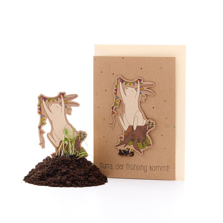 Una tarjeta de felicitación de primavera con un motivo de conejo y la inscripción "¡Hurra, llega la primavera!", junto a ella un conejo recortado con plántulas en la tierra.