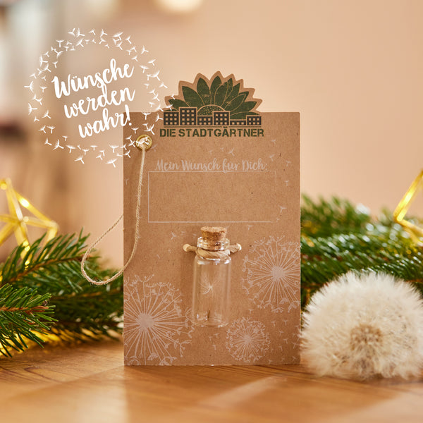 Tarjeta de felicitación de color marrón con la inscripción "Los jardineros de la ciudad", una botella de cristal y dientes de león decorativos junto a una rama de abeto; la inscripción "¡Los deseos se hacen realidad!".