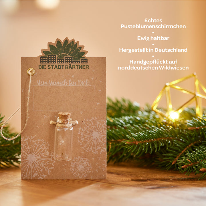Tarjeta con una botella y dientes de león, decorada con el logotipo de "Die Stadtgärtner" y un texto descriptivo. Ramas de abeto y luces de fondo.