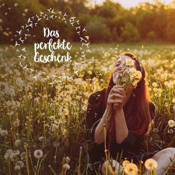 Mujer feliz sentada en un campo de dientes de león y oliendo flores con el texto "El regalo perfecto" encima.