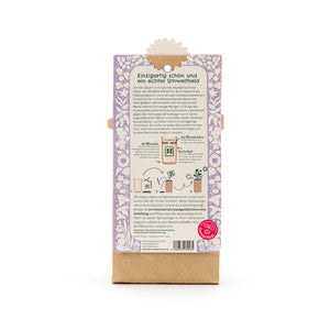 Parte posterior de un paquete con un patrón floral de color violeta, texto sobre el respeto al medio ambiente del árbol Kiri, instrucciones de plantación y el logotipo del Festival de Semillas en la parte inferior derecha.