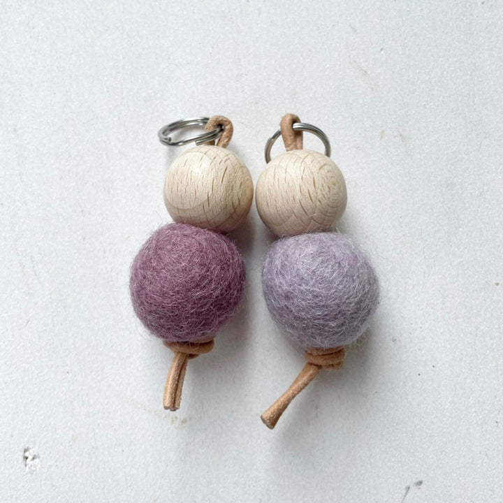 Dos llaveros hechos a mano con bolas de madera y bolas de fieltro de lana de color morado sobre fondo blanco.