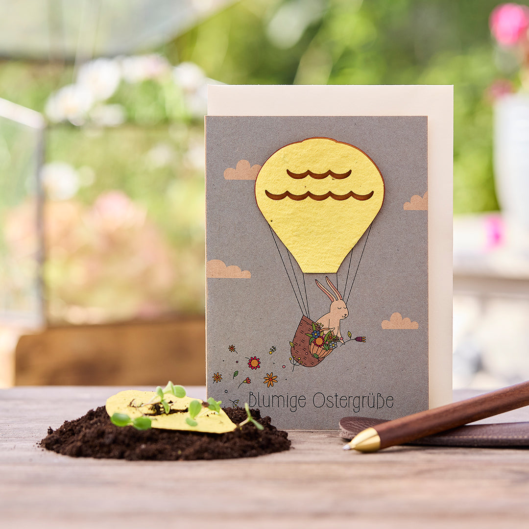 Tarjeta de Pascua con un conejito en un globo aerostático, al lado tierra con plantas germinando y un bolígrafo.