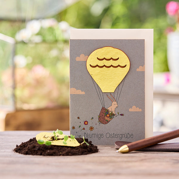 Tarjeta de Pascua con un conejito en un globo aerostático, al lado tierra con plantas germinando y un bolígrafo.