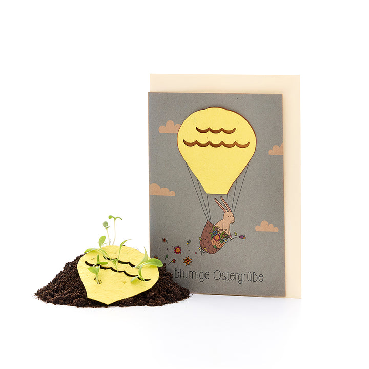 Tarjeta de Pascua con un conejito en un globo aerostático y papel de semillas en forma de corazón del que crecen pequeñas plantas en el suelo.