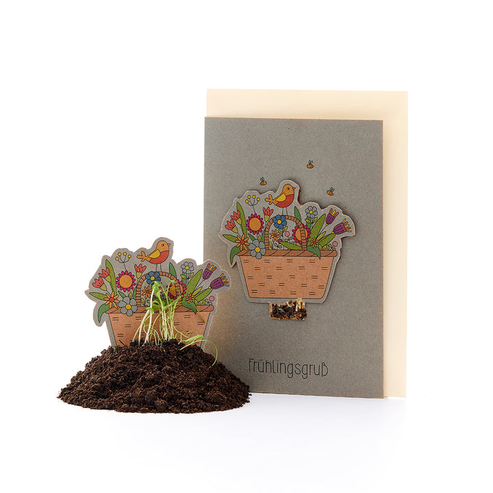 Tarjeta de felicitación con un motivo floral y un pájaro, además de una canasta de flores de papel en un montículo de tierra con plantas que brotan, texto "Saludos de primavera".