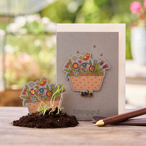 Tarjeta de felicitación con motivo primaveral, cesta de flores y pájaro, junto a ella un suelo con pequeñas plántulas y un bolígrafo sobre una mesa de madera.