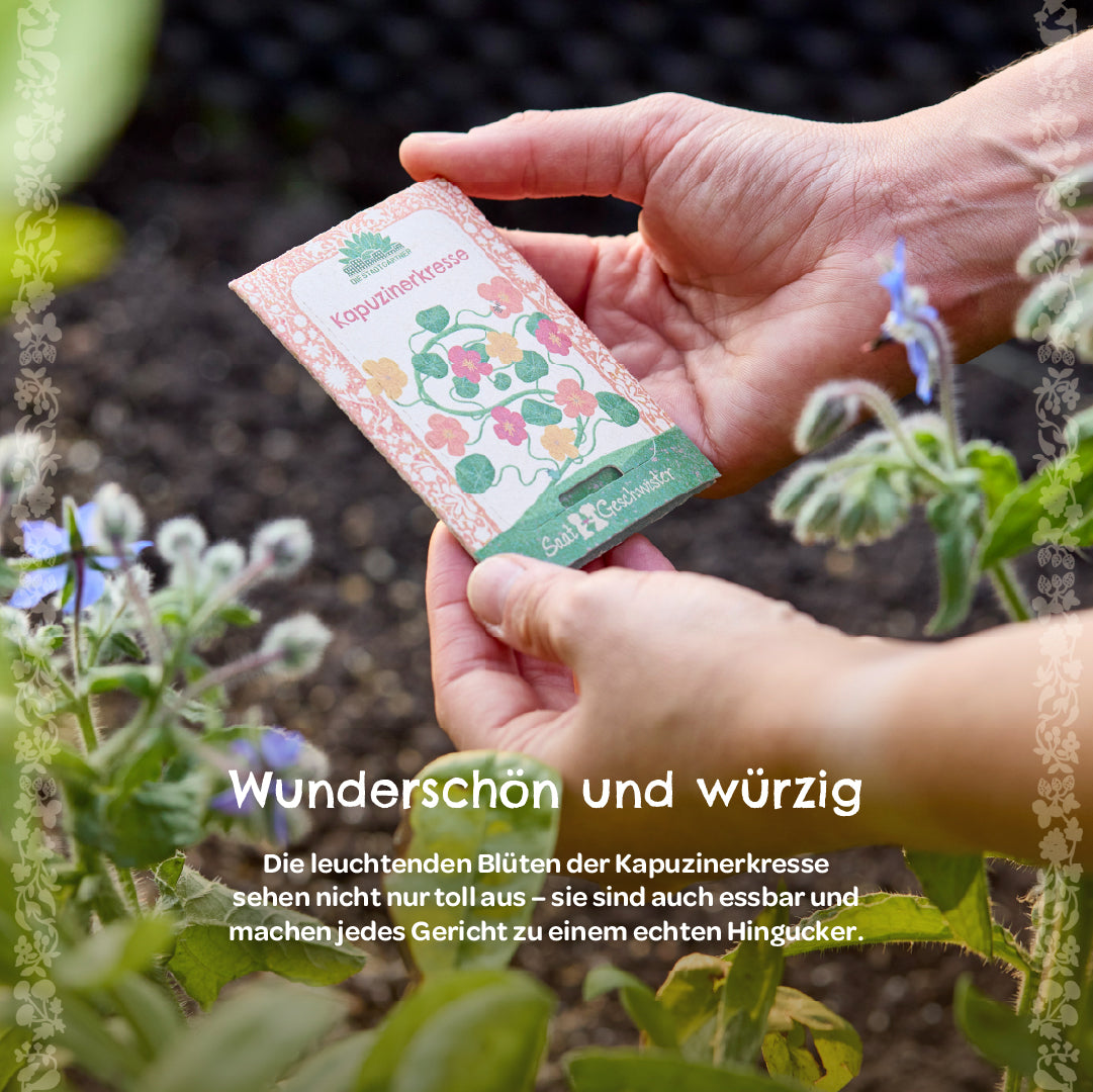Manos sosteniendo un paquete de semillas etiquetado como “capuchina” en el jardín, con plantas con flores al lado.