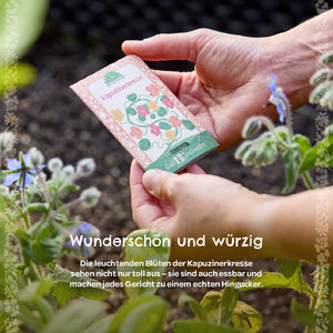 Manos sosteniendo un paquete de semillas etiquetado como “capuchina” en el jardín, con plantas con flores al lado.
