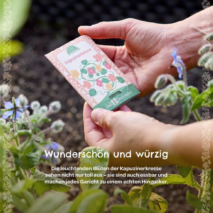 Manos sosteniendo un paquete de semillas etiquetado como “capuchina” en el jardín, con plantas con flores al lado.