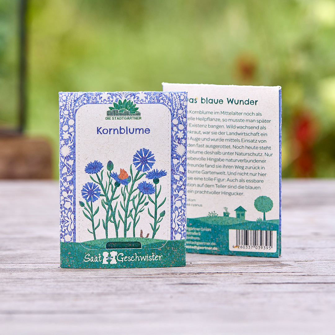 Dos paquetes de semillas de aciano de la marca "Saatgeschwister" sobre una mesa de madera, con una ilustración floral y una descripción del producto, y la naturaleza borrosa en el fondo.